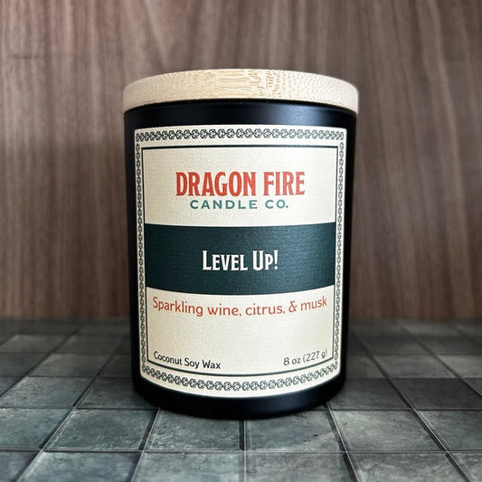 A Dragon Fire Candle Co. coconut soy wax candle in a black glass jar with a light wood lid, labeled 'Level Up!' on a dark tiled surface against a wood-paneled background.